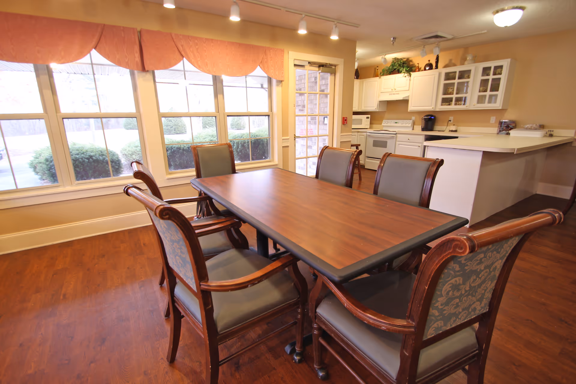 A dining area with a wooden table surrounded by six chairs with cushioned seats and backs. The room has large windows with peach-colored valances letting in natural light. In the background, there is a kitchen area with white cabinets, a stove, microwave, and a coffee maker. The floor is wooden, and the walls are painted beige.