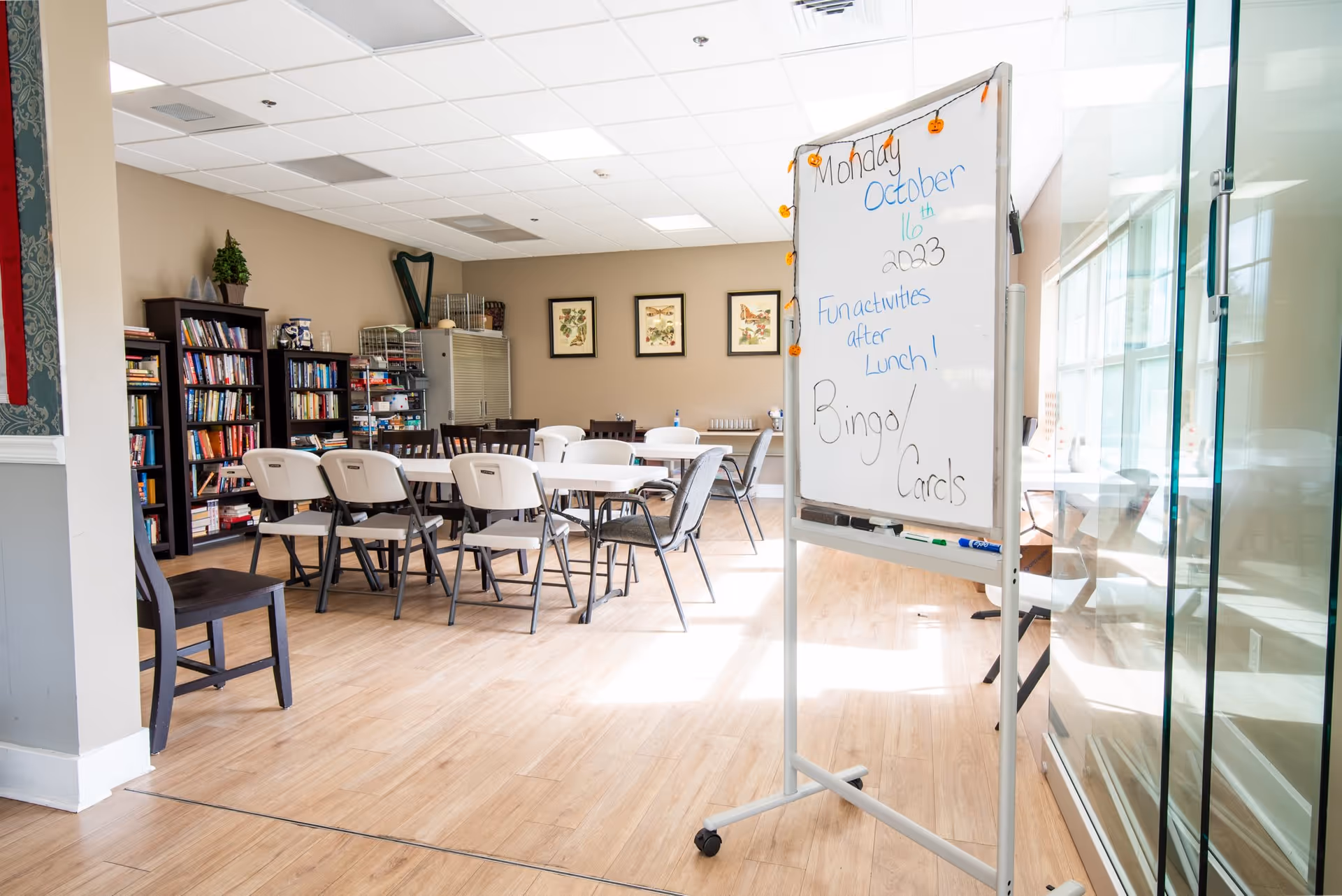 A bright and spacious activity room with several tables and chairs arranged for group activities. A whiteboard on wheels displays the date Monday, October 16th, 2023, and announces fun activities after lunch including Bingo and Cards. The room has bookshelves filled with books and games, framed botanical prints on the wall, and large windows letting in natural light.