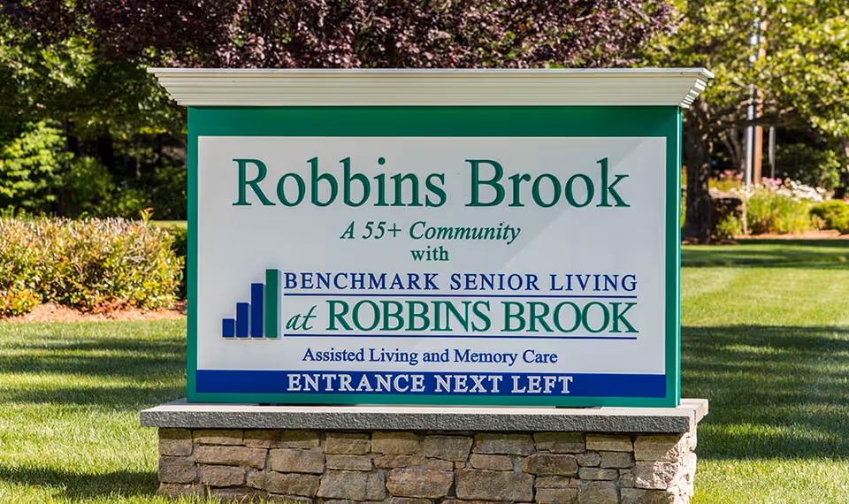 Outdoor sign for Robbins Brook, a 55+ community with Benchmark Senior Living at Robbins Brook, offering assisted living and memory care, with entrance indicated to the next left.