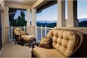 Covered balcony with cushioned patio chairs and an ottoman overlooking trees and distant hills at dusk.