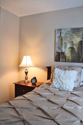 A cozy bedroom corner featuring a neatly made bed with a textured beige comforter and a decorative white pillow. Next to the bed is a wooden nightstand with a classic table lamp emitting warm light and a small round alarm clock. Above the bed hangs a framed artwork depicting a cityscape with tall buildings.