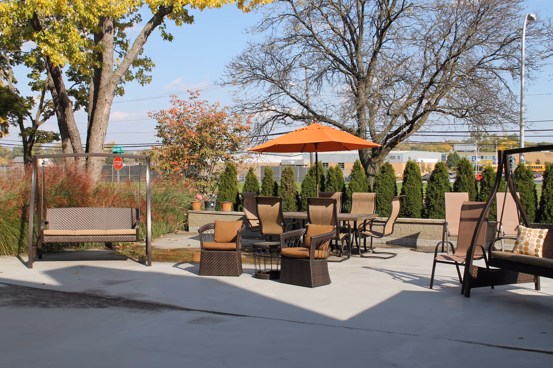 Outdoor patio area with brown wicker chairs and tables, an orange umbrella providing shade, two swing benches, and various trees and shrubs in the background under a clear blue sky.
