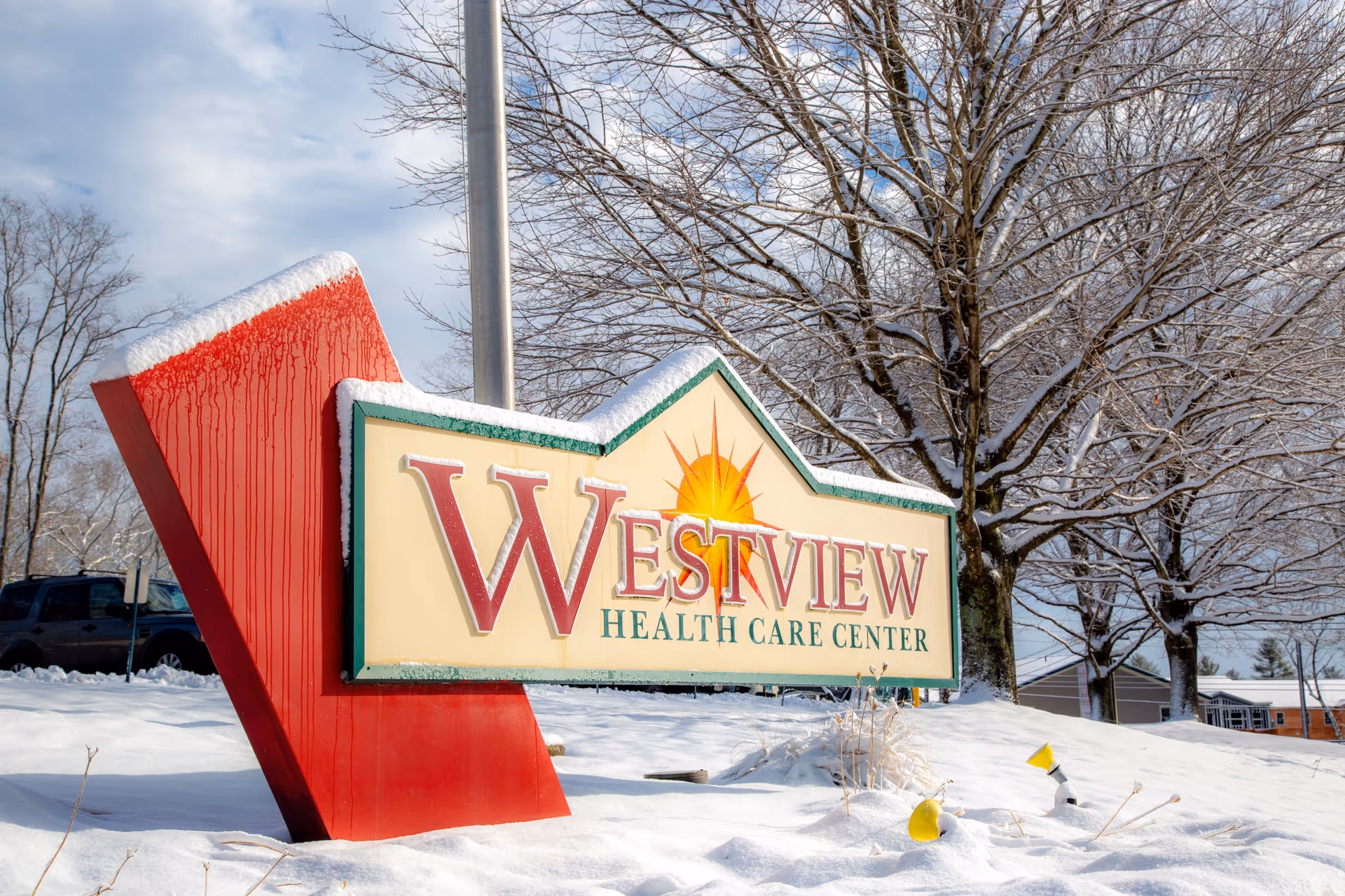 A large outdoor sign for Westview Health Care Center with a red base and a cream-colored panel featuring a sun graphic. The sign is surrounded by snow-covered ground and leafless trees under a partly cloudy sky.