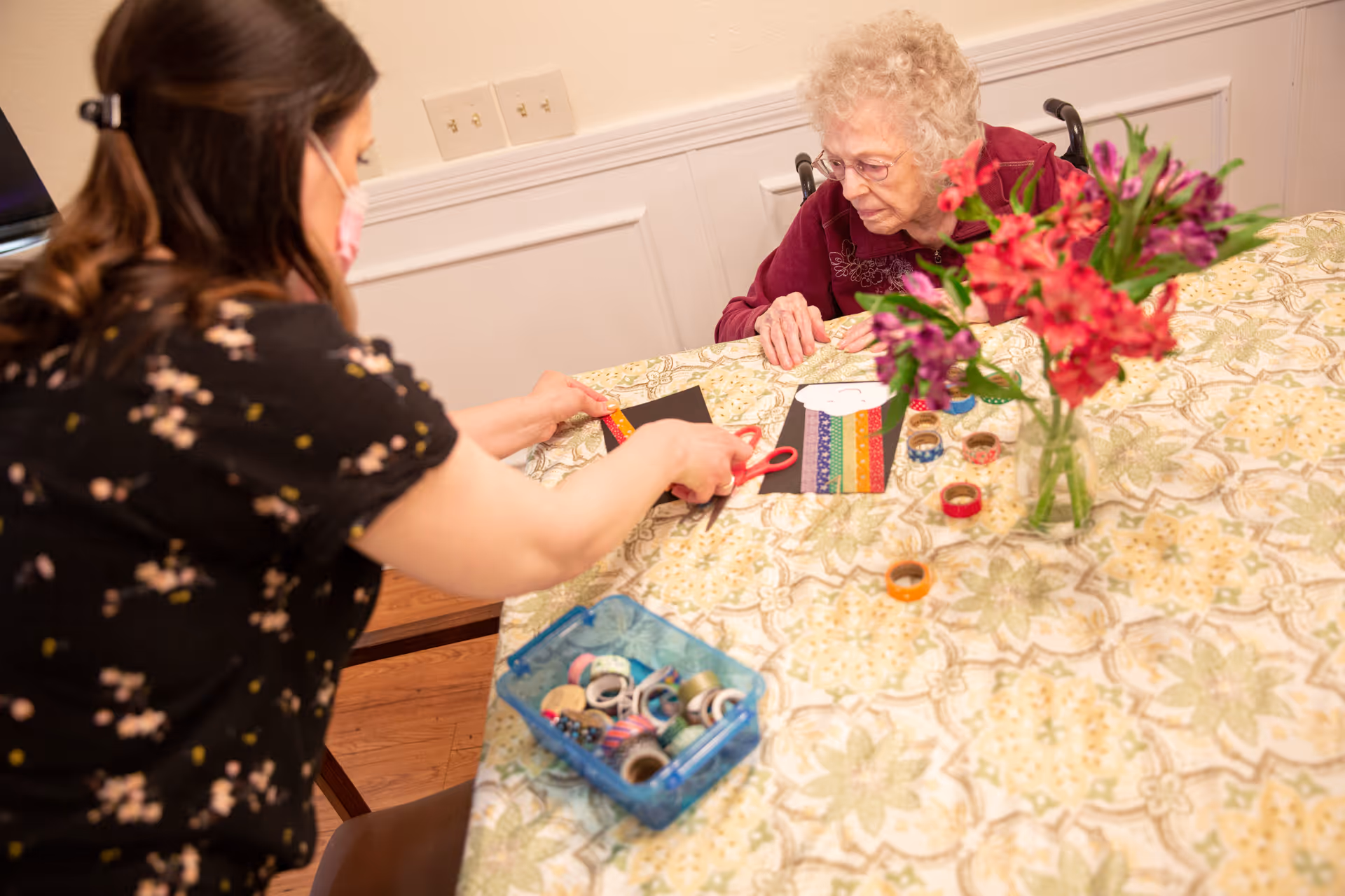 An elderly woman in a wheelchair and a caregiver wearing a mask are sitting at a table covered with a patterned tablecloth. The caregiver is cutting a piece of black paper with colorful tape on it, while the elderly woman watches. There is a vase with red and purple flowers and a container with various rolls of decorative tape on the table.