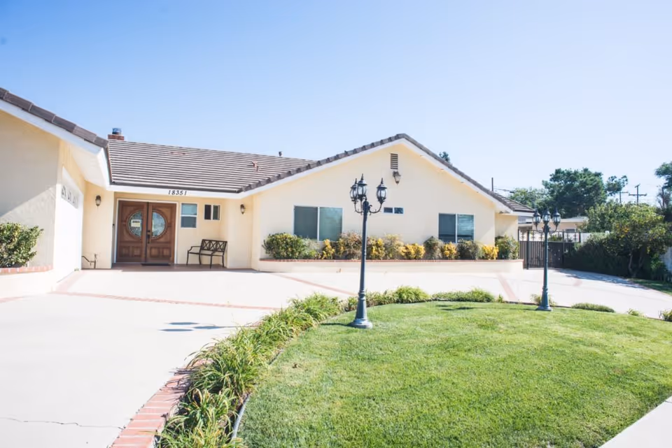 Exterior view of a single-story residential building with a tiled roof, beige walls, and a wooden double front door. The driveway and walkway are bordered by green grass and plants, with two black vintage-style lamp posts on the lawn. The sky is clear and blue.
