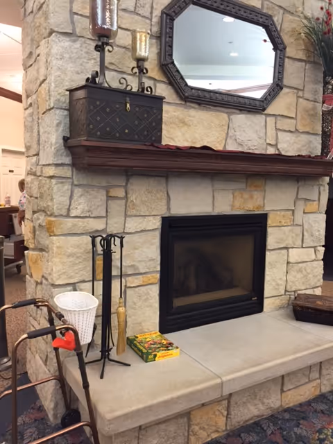 Stone fireplace with a wooden mantel holding decorative candle holders and a small chest. Above the mantel is a large octagonal mirror. In front of the fireplace are fireplace tools, a white basket, a walker, and a puzzle box on the hearth. The setting appears to be a cozy indoor common area.