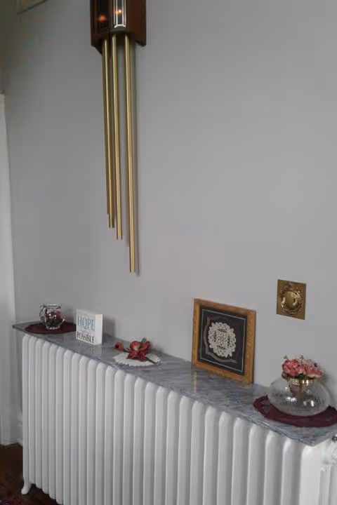 A white radiator with a marble top shelf decorated with small flower arrangements, a framed lace artwork, and a small sign that reads 'HOPE is possible'. Above the radiator hangs a wooden wind chime with long golden tubes. The wall behind is plain and light-colored.