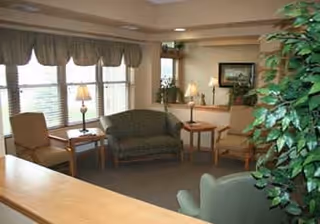 A cozy senior living community lounge area with several upholstered chairs and a loveseat arranged around wooden side tables. The room has large windows with valances, table lamps, framed artwork on the wall, and a large green plant in the foreground.