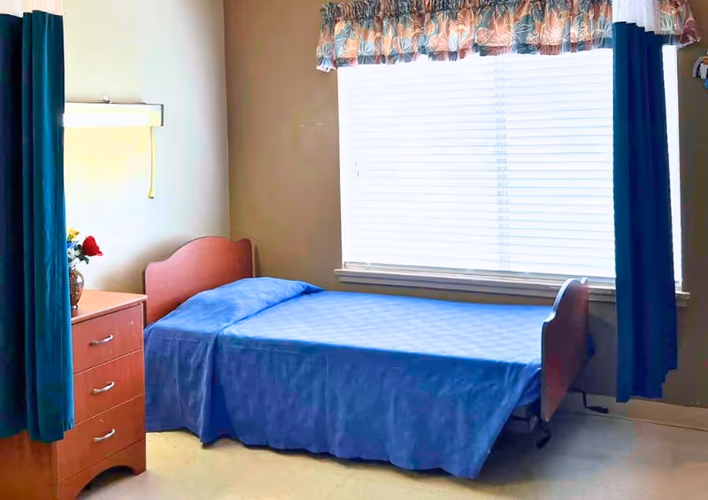 A simple bedroom with a single bed covered in a blue bedspread positioned against a wall with a large window. The window has white blinds and floral valance with dark blue curtains on either side. Next to the bed is a wooden nightstand with three drawers and a vase with flowers on top.