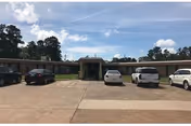 Front exterior view of a single-story nursing and rehabilitation facility named Woodland Manor Nursing & Rehabilitation, with a parking lot in front containing several parked cars under a partly cloudy sky.
