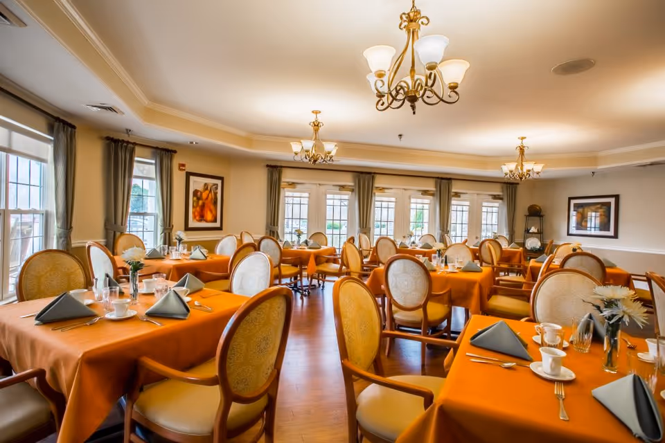 A bright and elegant dining room with multiple tables covered in orange tablecloths, each set with folded gray napkins, cups, glasses, and silverware. The room features large windows with green curtains, framed artwork on the walls, and ornate chandeliers hanging from the ceiling. The chairs have wooden frames with cushioned seats and backs.