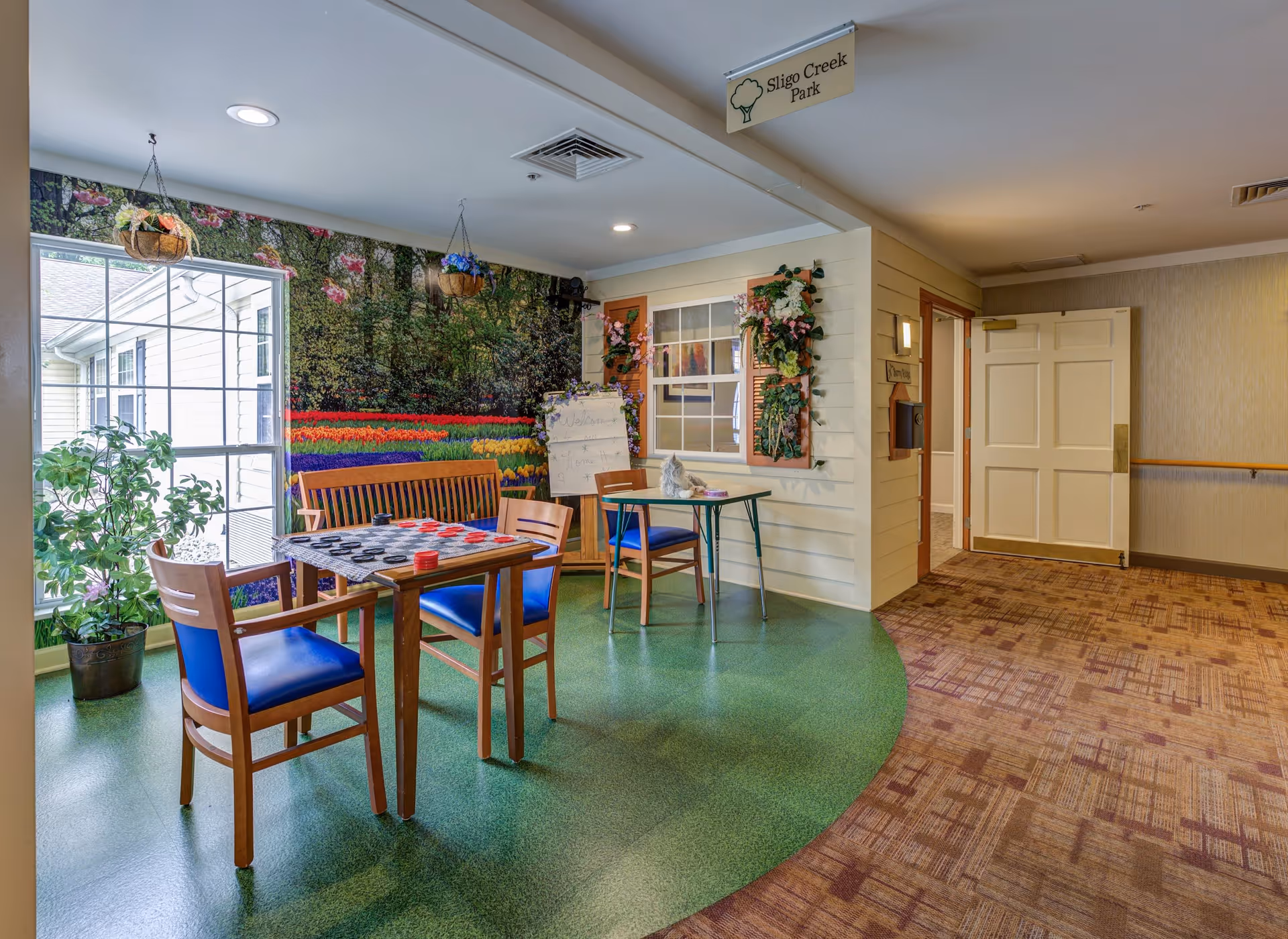 A cozy indoor seating area with two wooden tables and chairs with blue cushions. One table has a checkers game set up. The walls feature a large mural of a colorful flower garden and hanging flower baskets. A sign above reads 'Sligo Creek Park'. There is a window and a door leading to another room, with carpeted flooring transitioning to green linoleum.