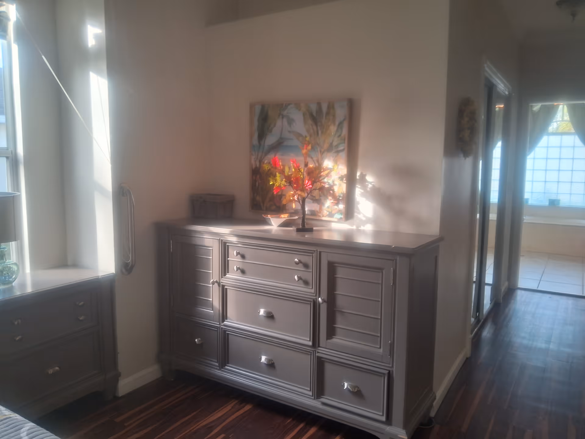 A softly lit interior hallway area with a gray wooden dresser featuring multiple drawers and cabinets. On top of the dresser is a decorative arrangement of colorful artificial flowers and a framed painting of plants on the wall behind it. To the left, there is a window allowing natural light to enter, and to the right, a hallway leads to another room with large windows covered by sheer curtains.