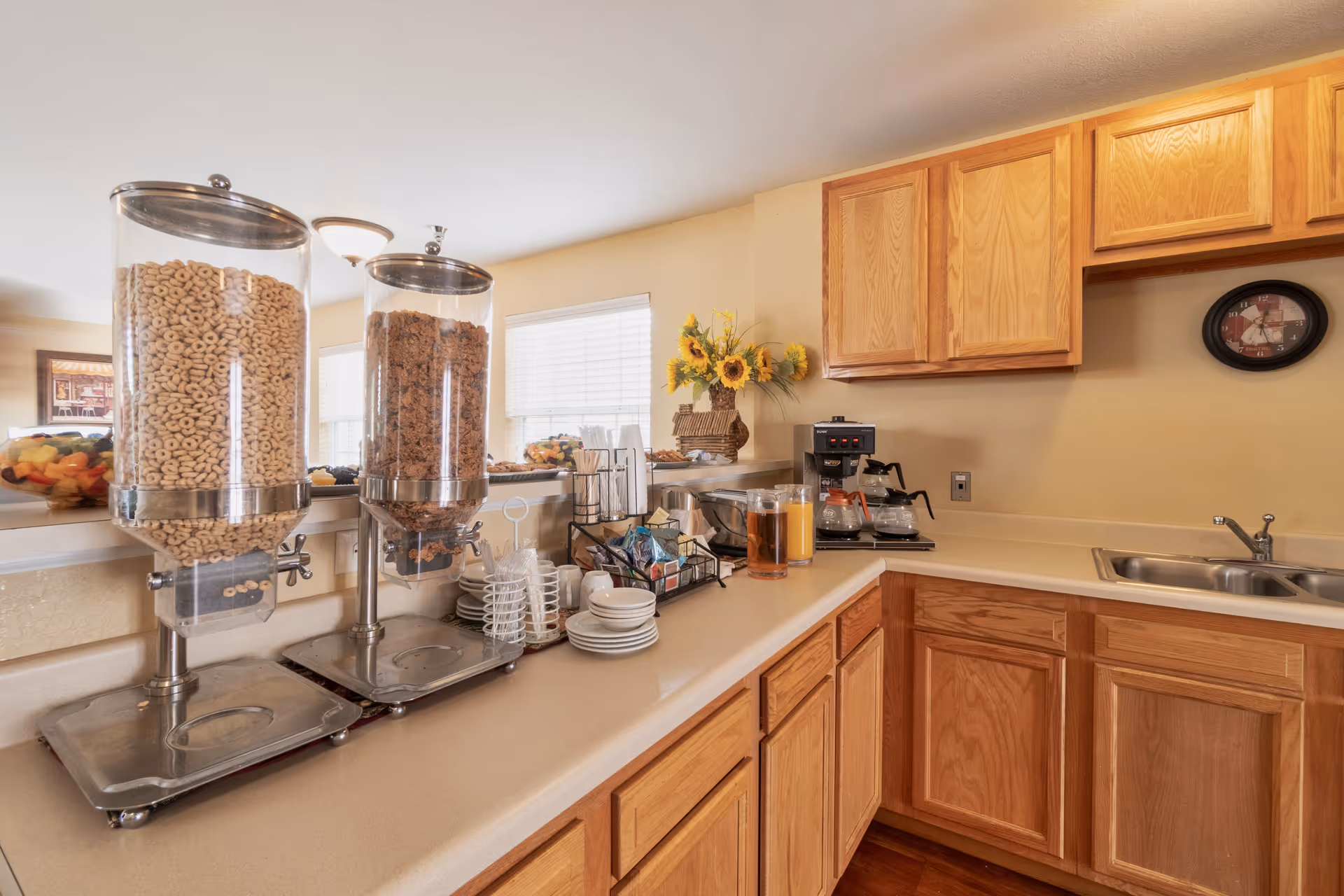 A kitchen area with wooden cabinets and a countertop featuring two cereal dispensers filled with Cheerios and granola, a coffee maker with two coffee pots, glasses of juice, plates, cups, and a basket with sunflowers. There is a double sink and a wall clock above the counter.