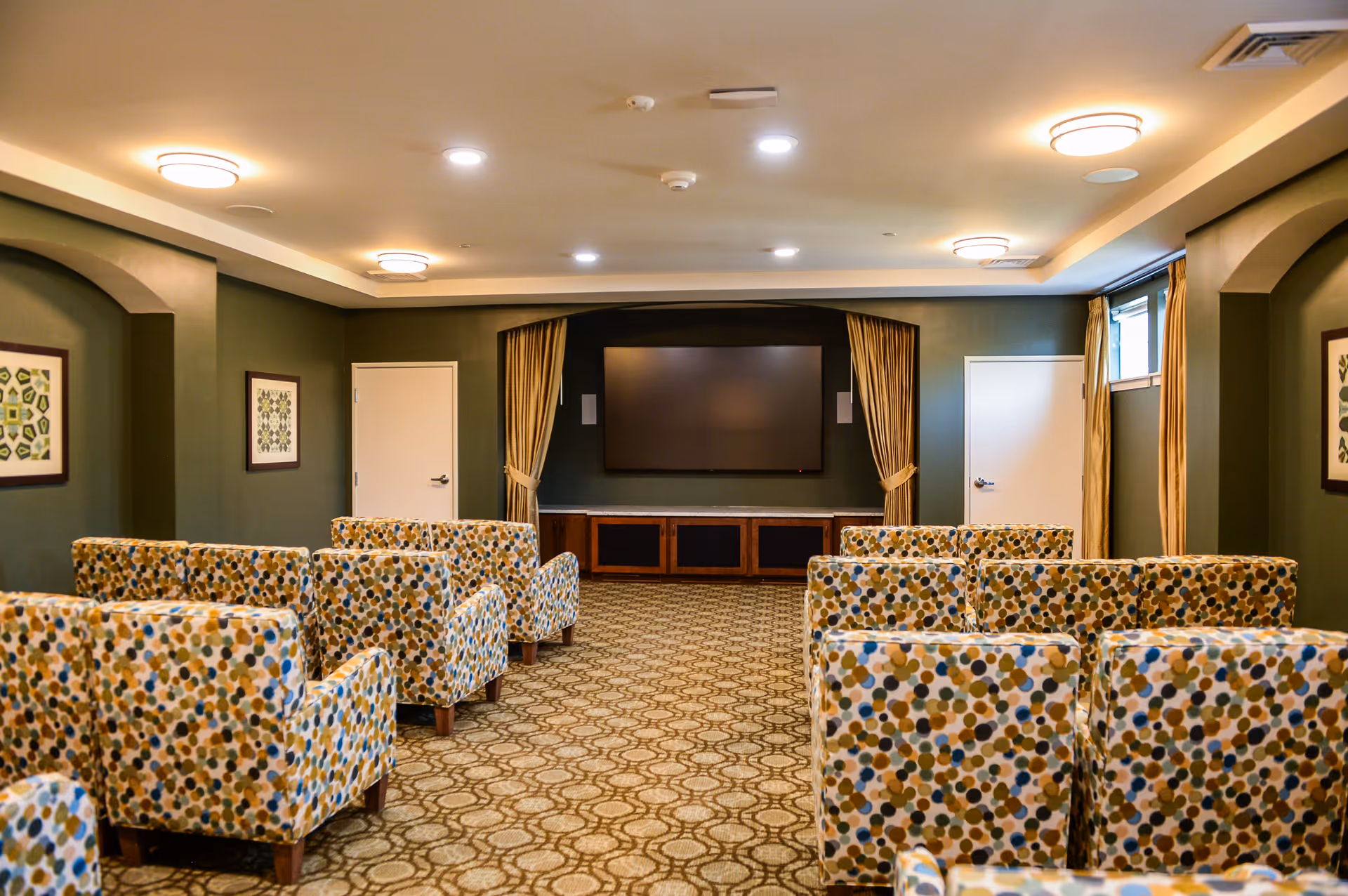A cozy media room with patterned armchairs arranged in rows facing a large flat-screen TV mounted on a dark green wall. The room has a patterned carpet, framed artwork on the walls, ceiling lights, and two doors on either side of the TV with gold curtains framing the TV area.