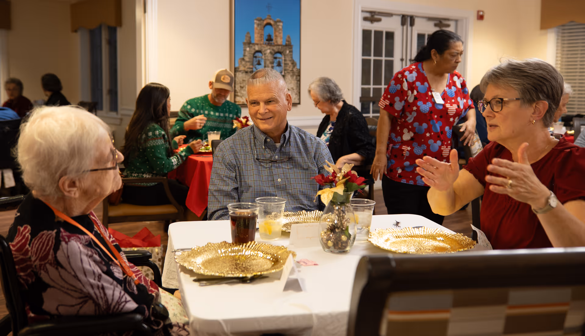 Several elderly people and a staff member are gathered in a dining area. Three seniors are seated at a table with gold decorative plates and drinks, engaging in conversation. In the background, other residents are seated at tables with red tablecloths, and a staff member in a red patterned shirt is walking by. The room has warm lighting and a picture of a bell tower on the wall.