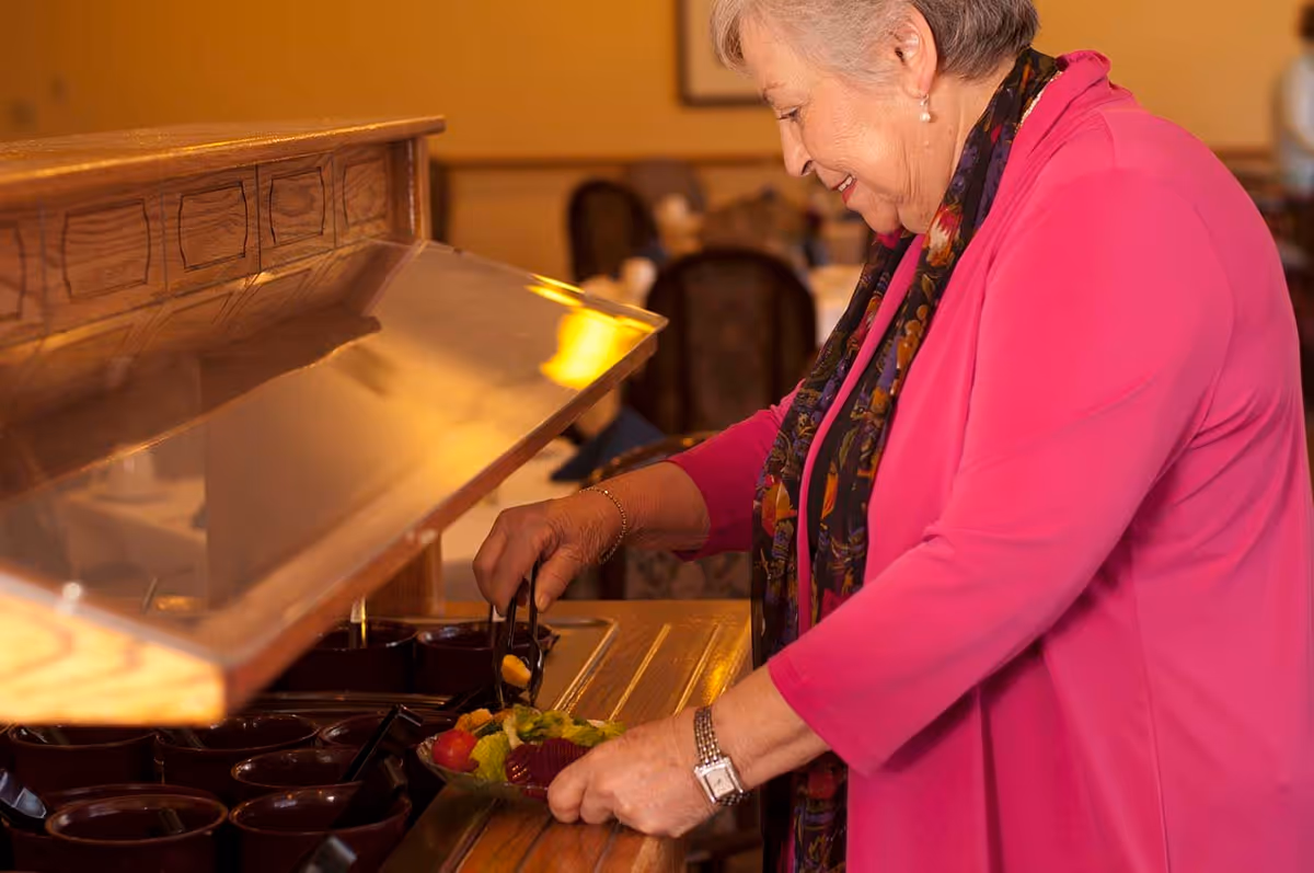 An elderly woman wearing a bright pink jacket and a patterned scarf is serving herself fruit salad from a buffet in a dining area with wooden furniture and warm lighting.