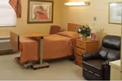 A nursing home bedroom with a single bed covered in an orange bedspread, a wooden bedside table with a flower vase, a brown leather recliner chair, a window with blinds and a valance, and framed artwork on the walls.