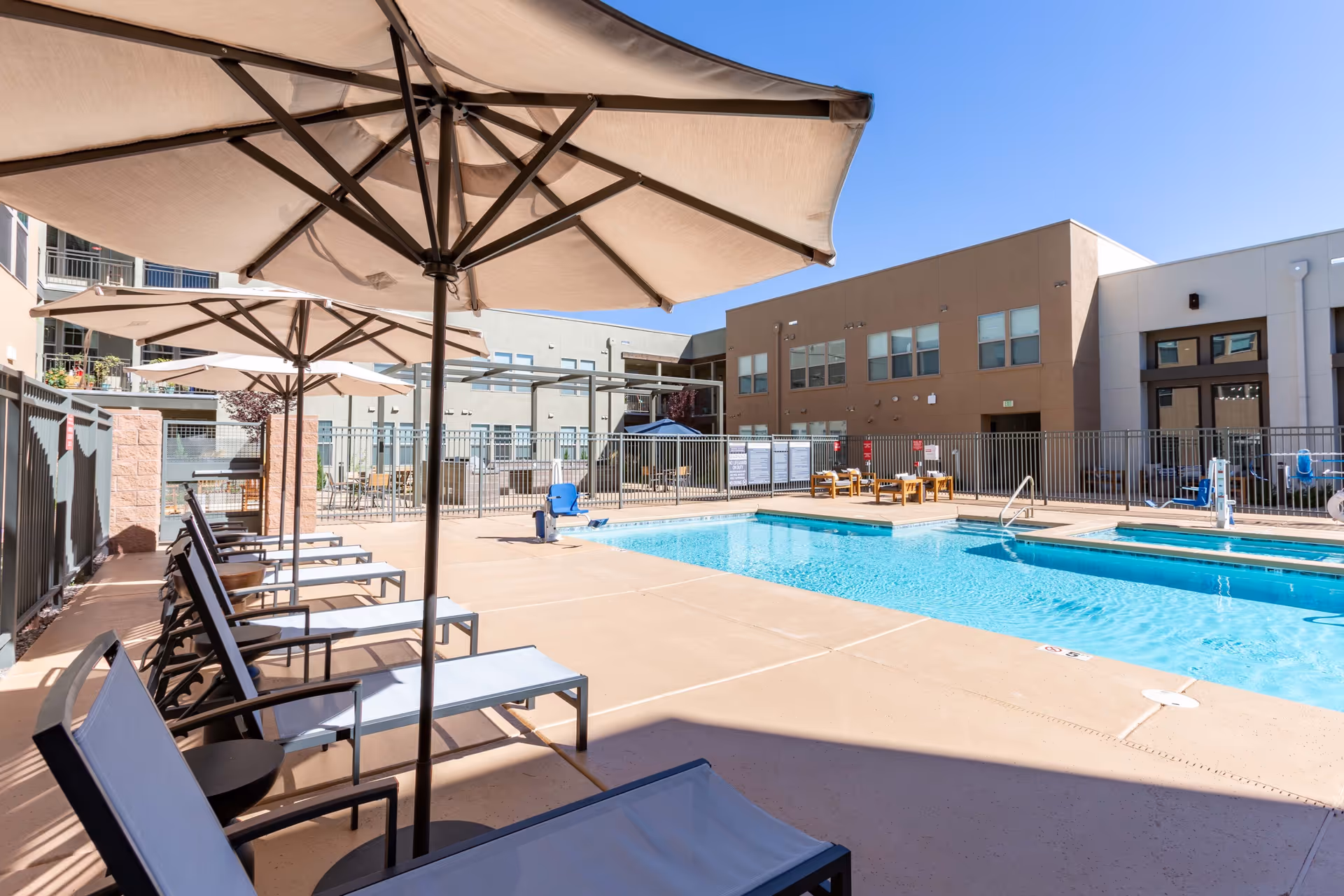 Outdoor swimming pool area at Overture Andalucia with lounge chairs and large beige umbrellas along the poolside, surrounded by a fence and multi-story buildings under a clear blue sky.