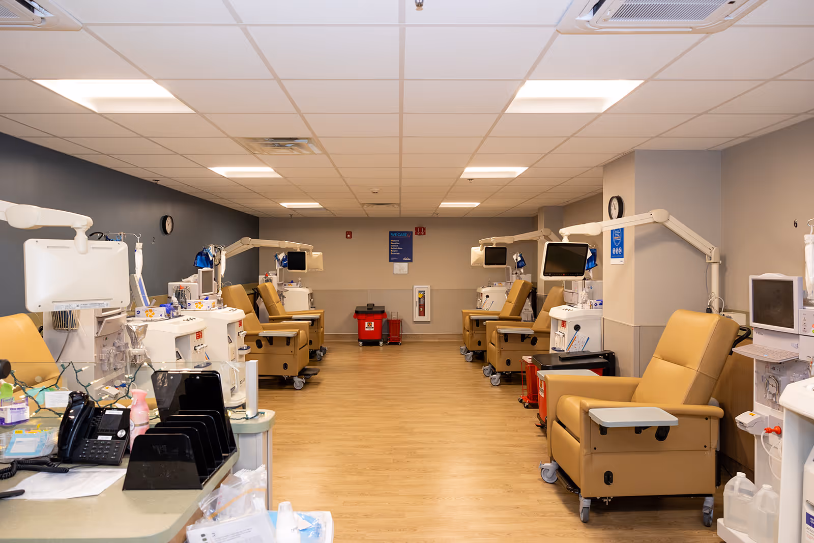 A medical treatment room with multiple beige recliner chairs arranged in two rows facing each other. Each chair is equipped with medical machines and monitors, likely for dialysis or infusion therapy. The room has a light wood floor, a drop ceiling with fluorescent lights, and a blue and beige color scheme on the walls. There is a counter with office supplies and a telephone in the foreground on the left side.
