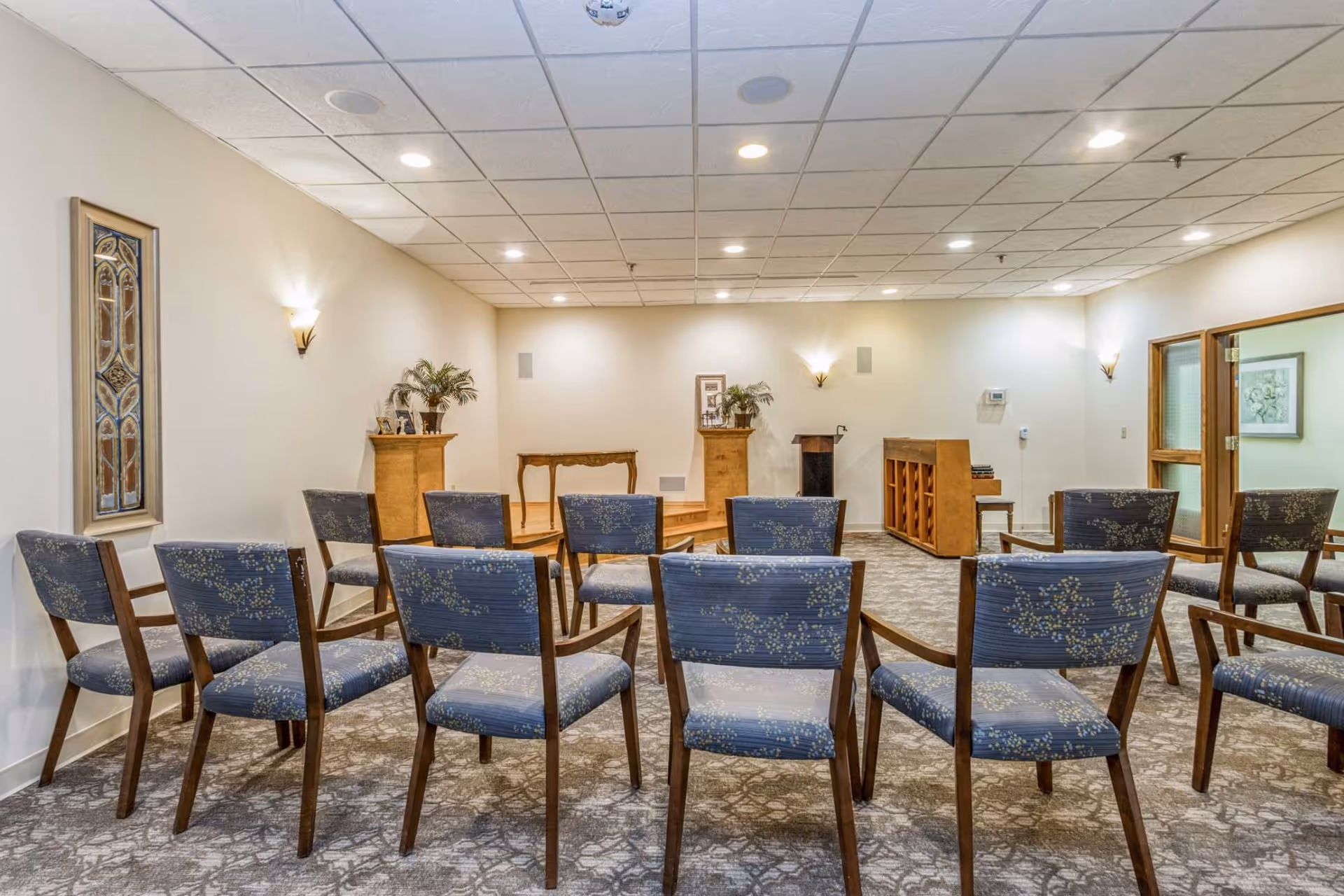 Small chapel-style meeting room with rows of upholstered chairs facing a podium and piano.