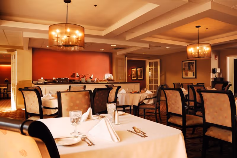 A warmly lit dining room with several tables covered in white tablecloths, each set with glassware, napkins, and silverware. The room features cushioned chairs with patterned upholstery, two large pendant lights hanging from the ceiling, and a red accent wall in the background with a buffet area.