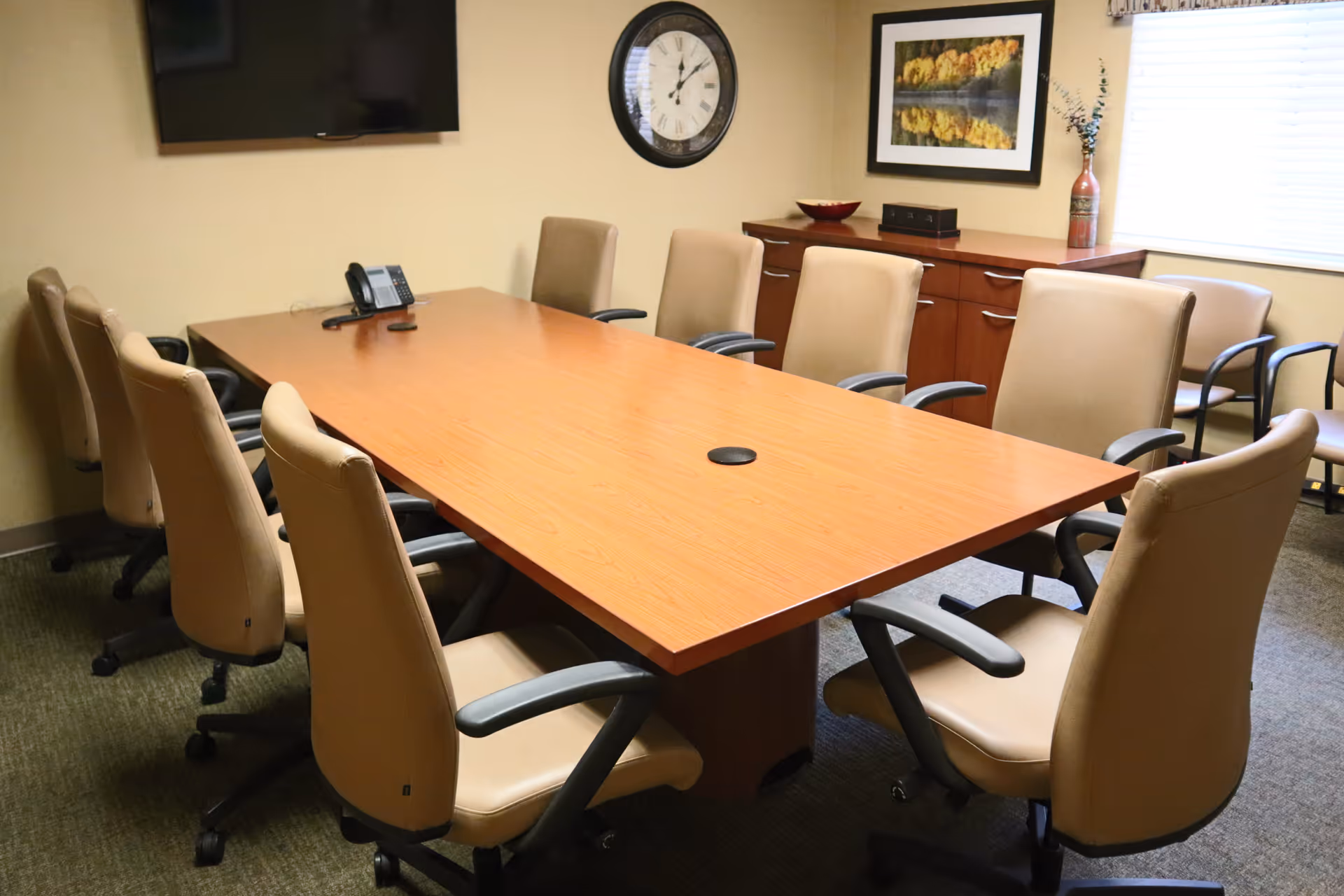 A conference room with a large rectangular wooden table surrounded by beige office chairs on wheels. On the wall, there is a large clock, a flat-screen TV, and a framed floral picture. A wooden cabinet with decorative items is positioned under a window with blinds.