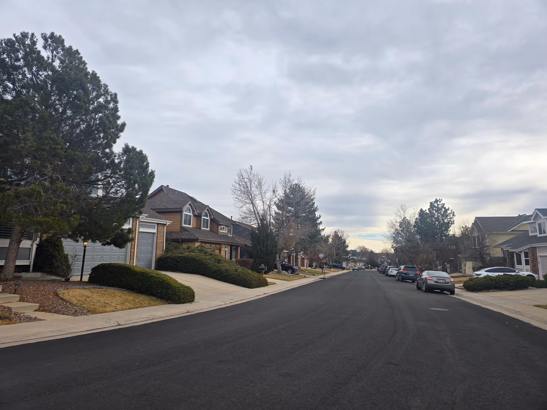 A suburban residential street with houses on both sides, some trees, parked cars along the curb, and a cloudy sky overhead.