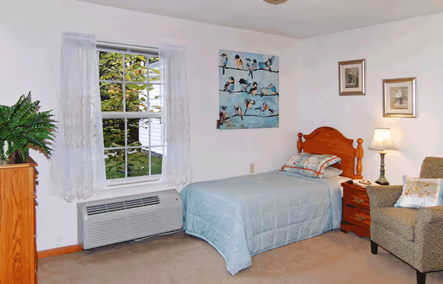 A cozy single-occupancy bedroom with a twin bed, nightstand and lamp, armchair, window with lace curtains, and wall art.