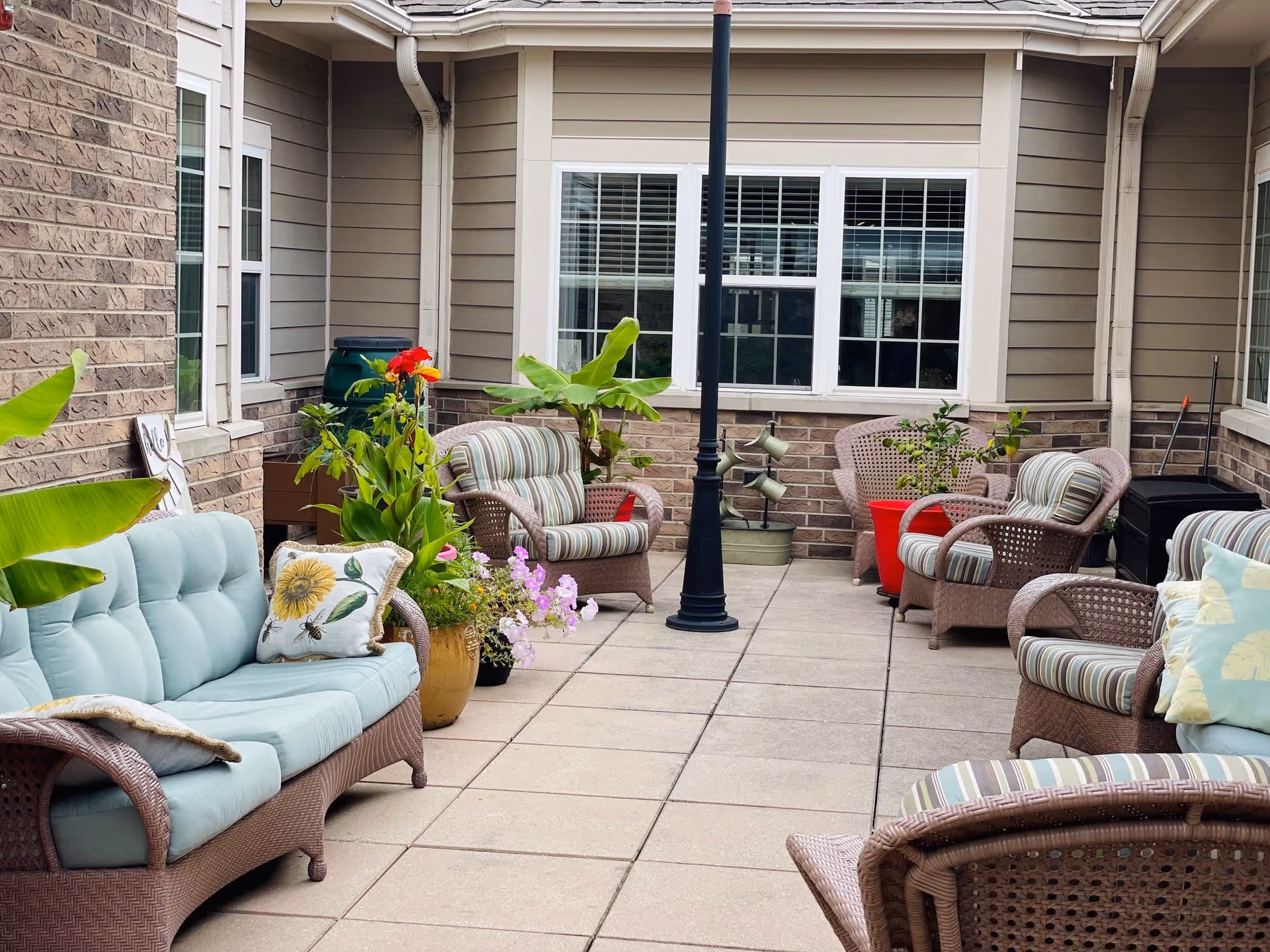 Outdoor patio area with cushioned wicker chairs and sofas arranged around a central lamp post. Several potted plants with green leaves and colorful flowers decorate the space. The patio is surrounded by the exterior walls of a building with windows and beige siding.
