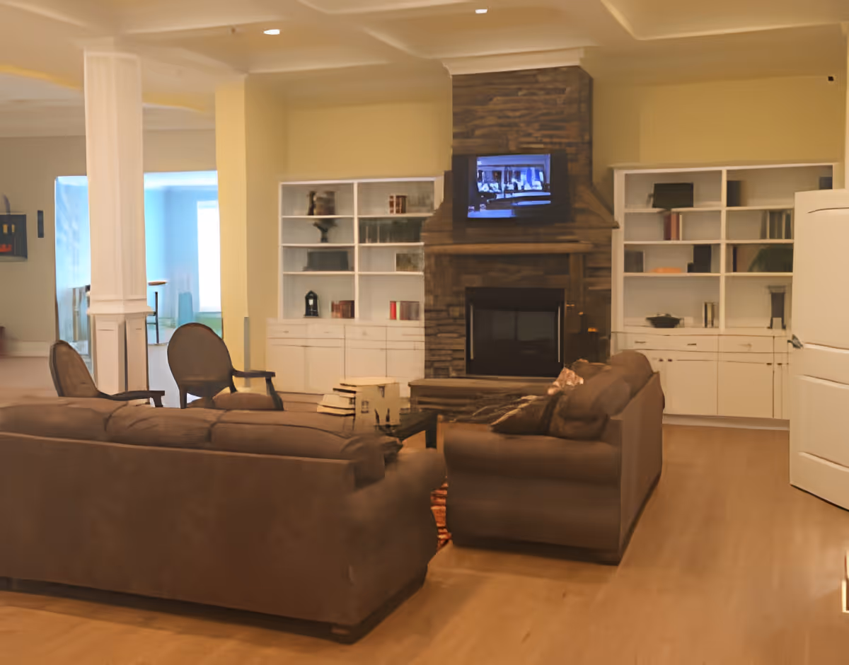A cozy living room with brown sofas and chairs arranged around a stone fireplace with a TV mounted above it. There are white built-in shelves on either side of the fireplace, some with books and decorative items. The room has light-colored walls and wooden flooring.