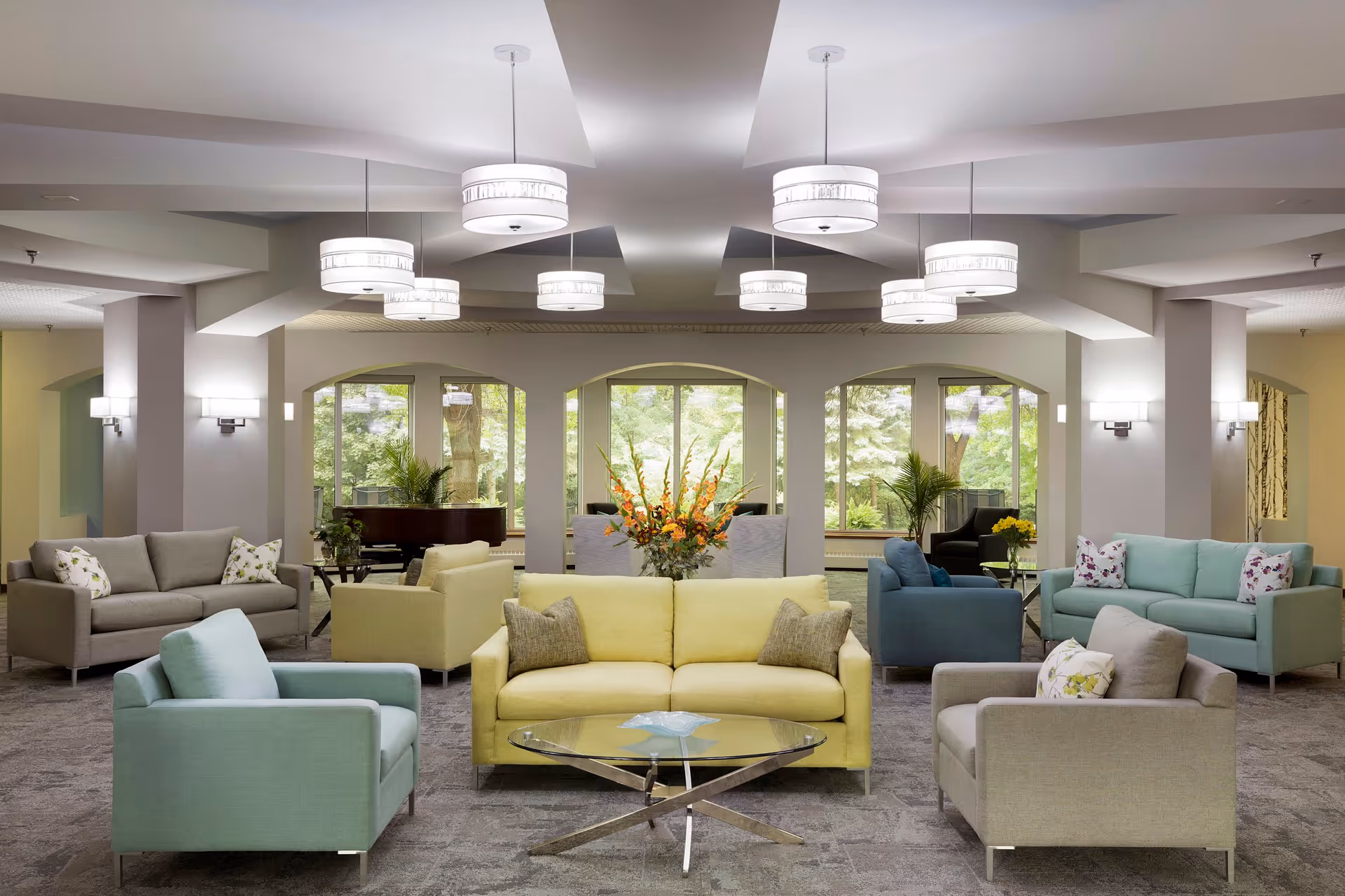 A spacious and well-lit senior living facility lounge area with multiple sofas and armchairs in pastel colors arranged around a glass coffee table. Large windows in the background reveal greenery outside, and modern ceiling lights illuminate the room. There are decorative plants and a floral arrangement on a table in the center.
