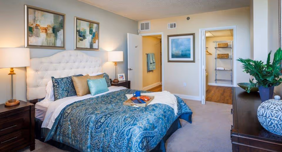 A well-lit bedroom with a large bed featuring a tufted white headboard and blue paisley bedding. Two bedside tables with lamps flank the bed. The room has light-colored walls adorned with abstract artwork. An open door reveals a walk-in closet with shelves and hanging space. A dresser with a plant and decorative items is positioned near a window with blinds.