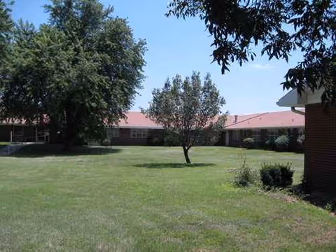 Single-story brick building with a red roof, green lawn, and several trees in front.