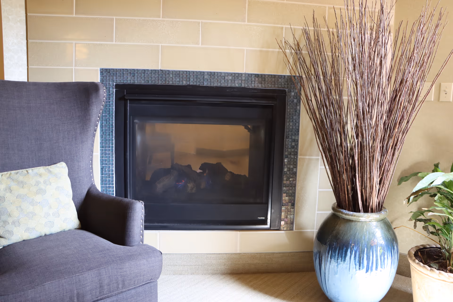 A cozy corner featuring a dark gray upholstered armchair with a light patterned cushion, positioned next to a modern fireplace with a black frame and small mosaic tiles surrounding it. To the right of the fireplace is a large ceramic vase with tall dried branches, and a smaller potted green plant is partially visible.