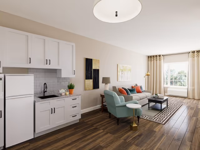 Open living room with a small kitchenette, sofa, armchair, coffee table, and a large window with curtains letting in natural light.
