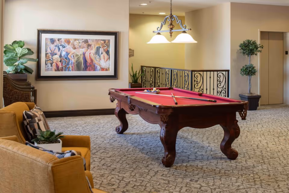 Indoor recreation area with a red-felt pool table, seating, artwork on the wall, and potted plants.