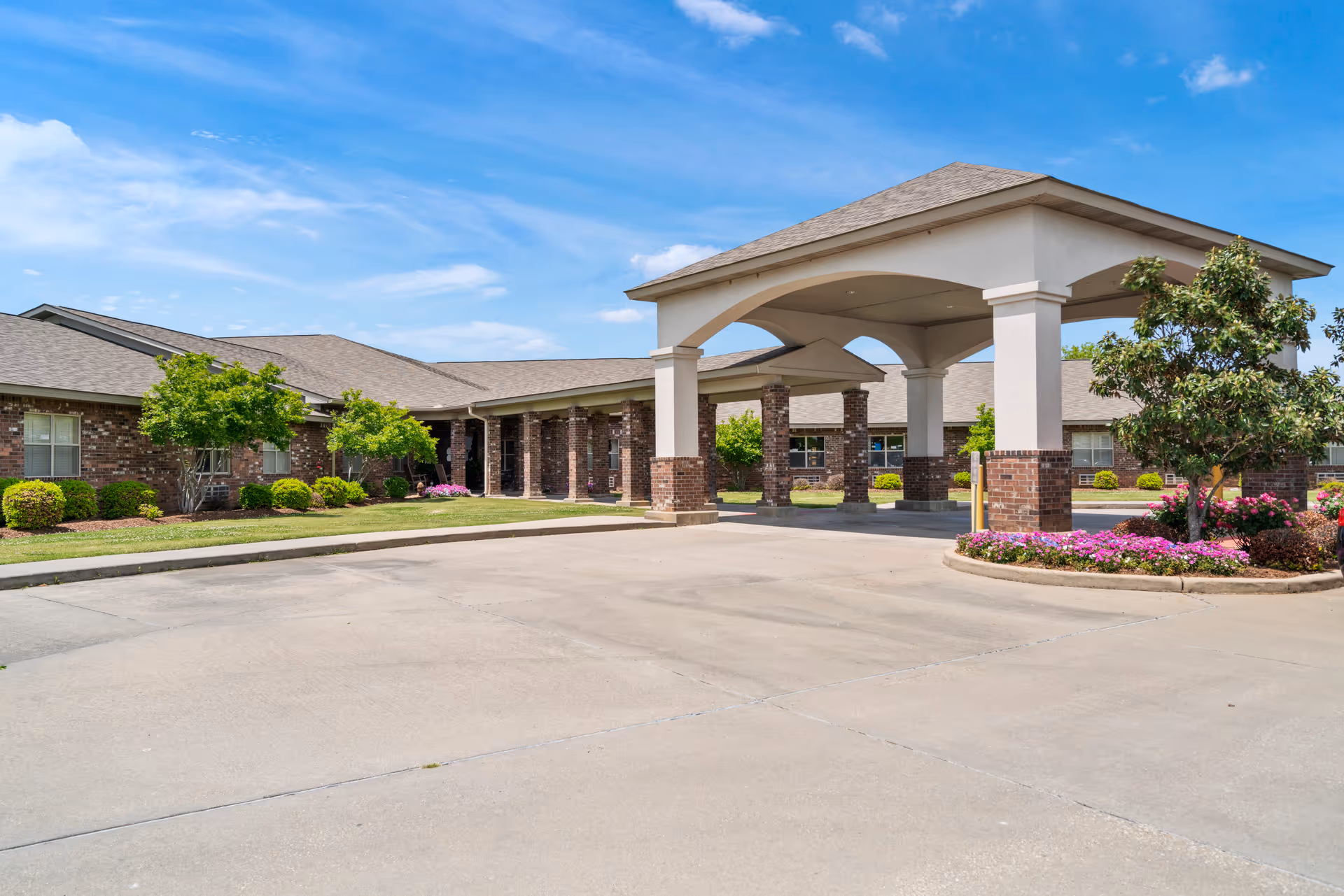 Exterior view of Heritage Manor Stratmore showing a covered entrance with brick pillars, a driveway, landscaped bushes, trees, and flowers under a blue sky with some clouds.