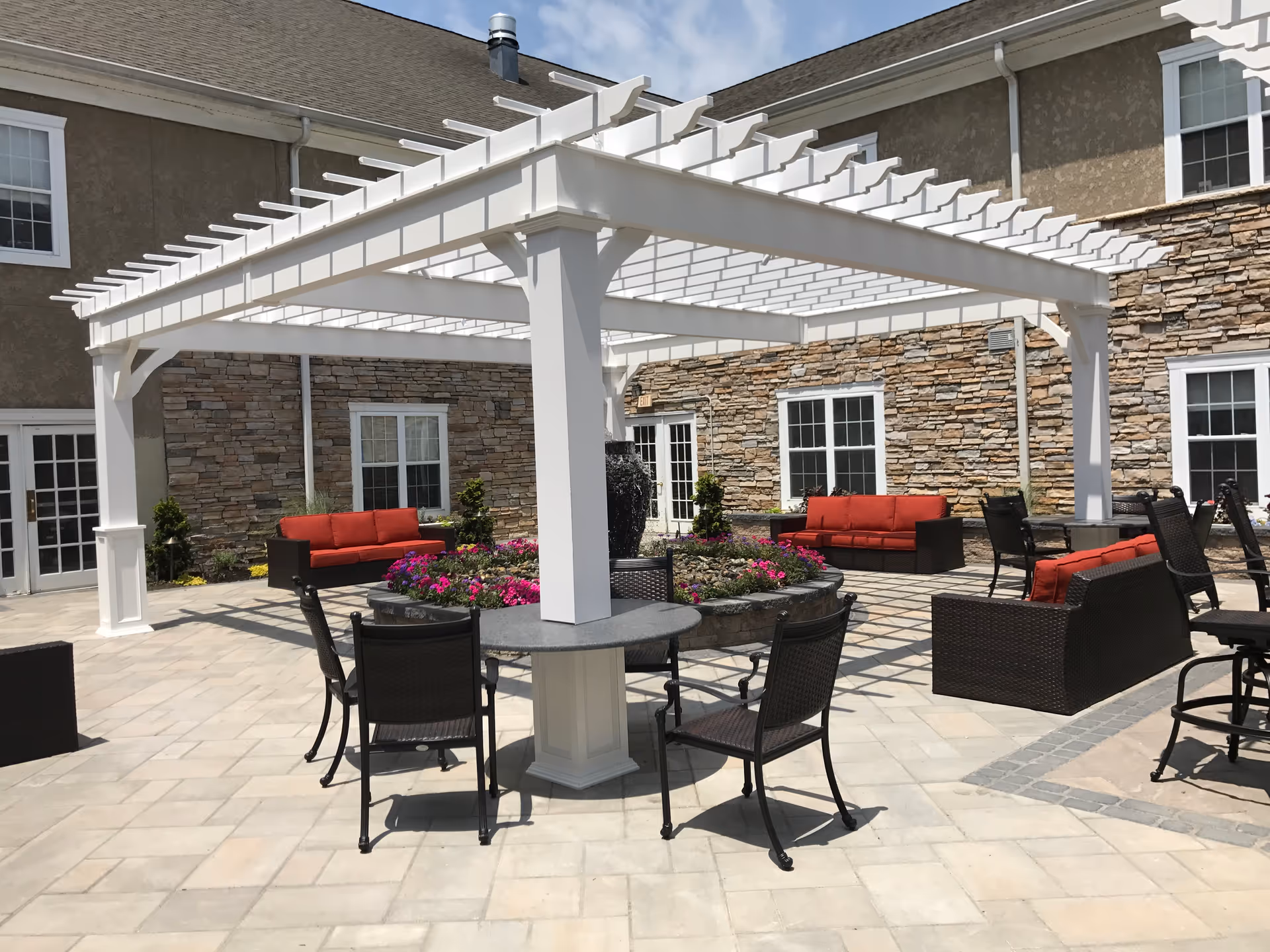 Outdoor patio area with a white pergola providing partial shade over a round table with four chairs. Surrounding the pergola are several seating areas with dark wicker furniture and red cushions. In the center of the patio is a circular flower bed with colorful flowers and a water fountain. The background shows a building with stone and beige stucco walls and multiple windows.