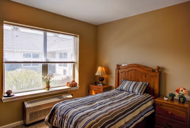 A cozy bedroom with a single wooden bed featuring a striped comforter and pillow. There is a wooden nightstand with a lamp and alarm clock next to the bed. Another wooden dresser with small potted plants and flowers is on the right side. A large window with blinds lets in natural light, and a heating/cooling unit is positioned below the window.