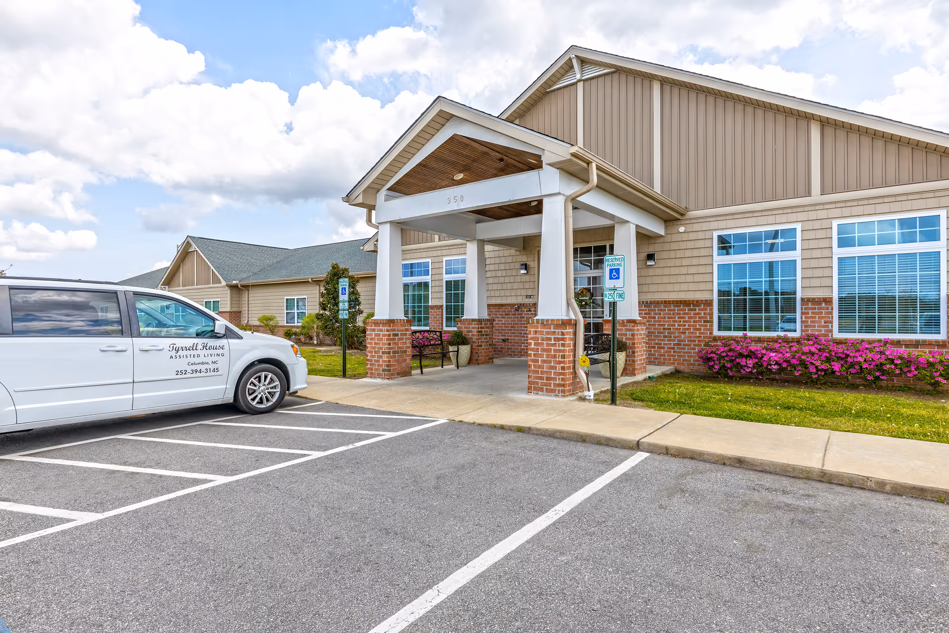 Front exterior view of Tyrrell House assisted living facility showing the entrance with a covered porch supported by white columns and brick bases. A white van with Tyrrell House branding is parked in a handicapped parking space near the entrance. The building has beige siding, large windows with white trim, and a flower bed with pink flowers along the front. The sky is partly cloudy.