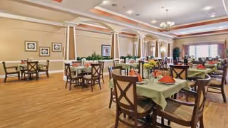 Spacious dining room with multiple tables covered in green and orange tablecloths, set with plates and napkins. The room features wooden floors, beige walls with framed artwork, large windows with curtains, and ceiling lights including chandeliers.