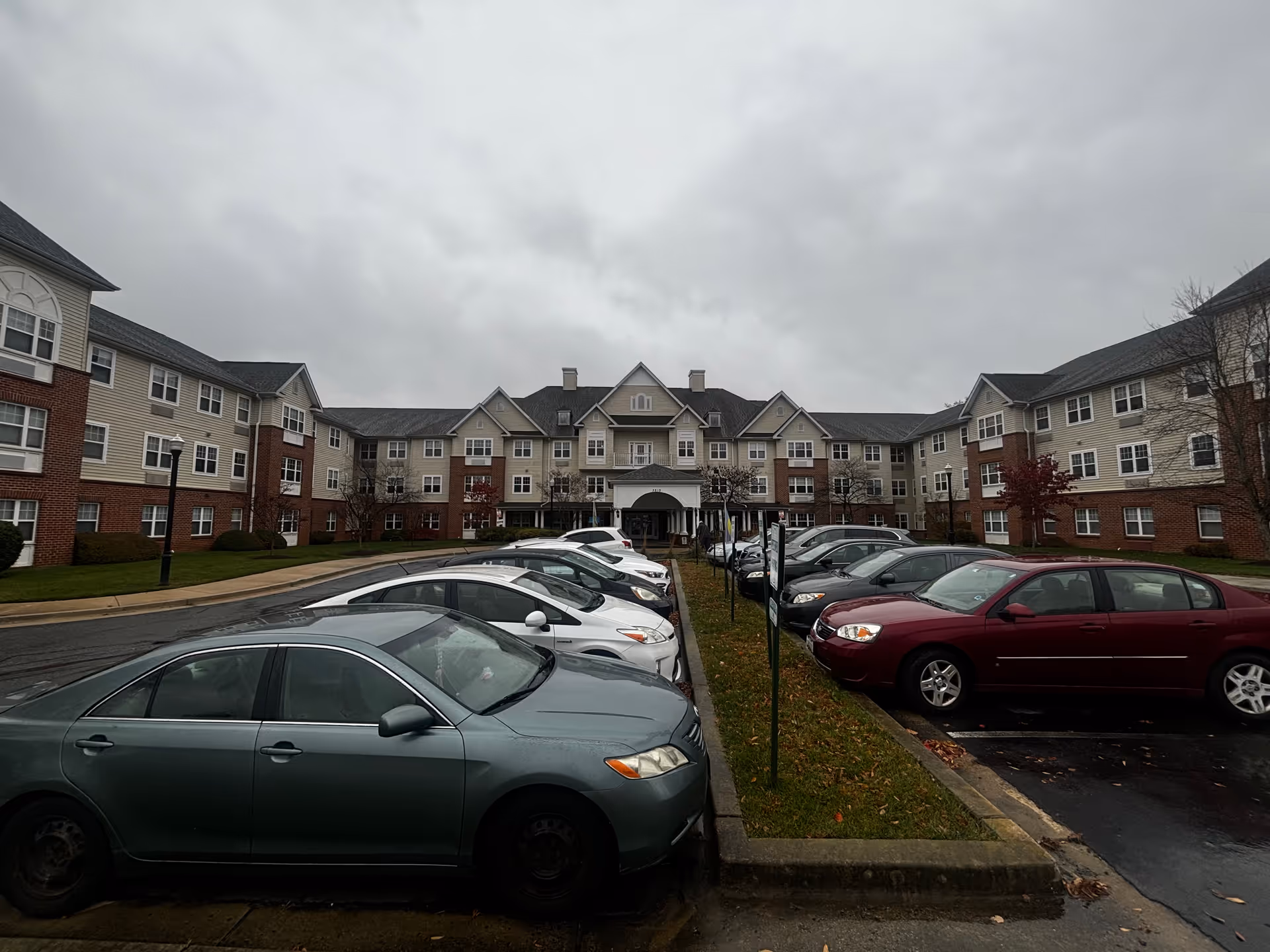 Exterior view of a large multi-story senior living facility named Grace House on a cloudy day, with a parking lot filled with cars in the foreground and a driveway leading to the main entrance.