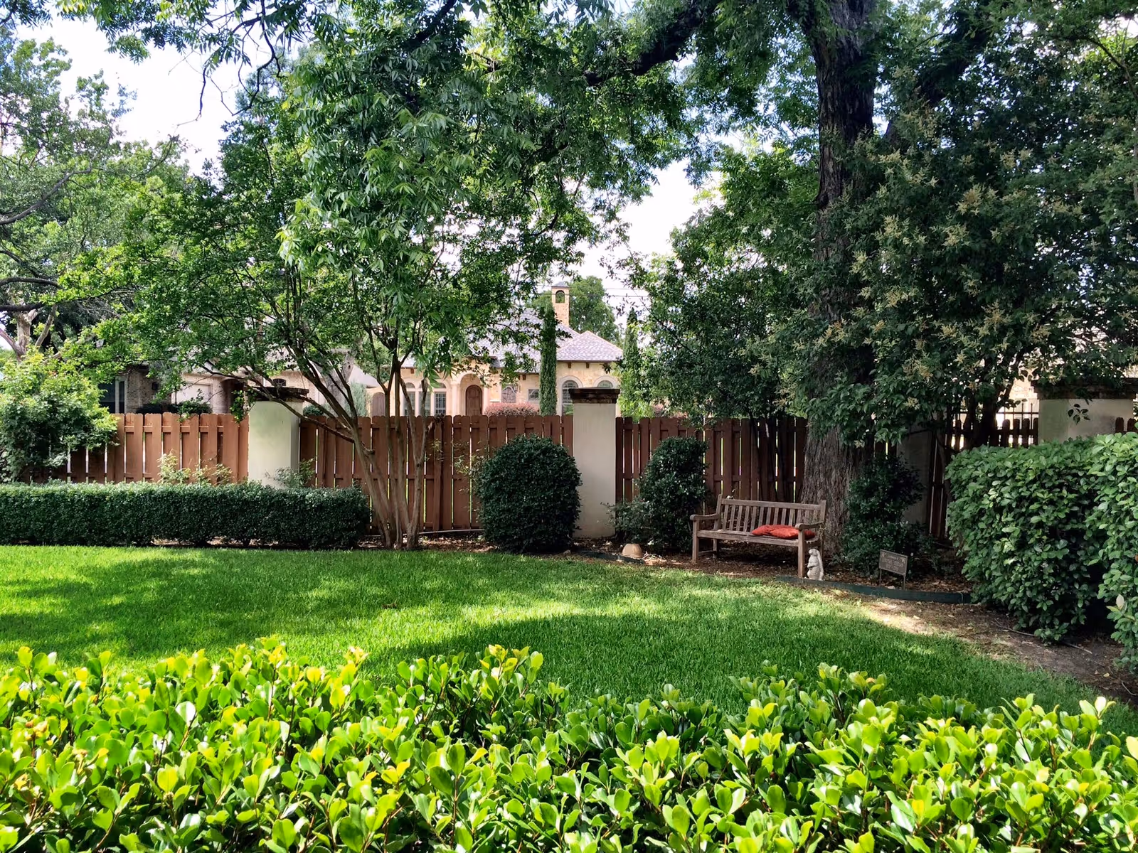 A lush green garden area with neatly trimmed bushes and a wooden bench with a red cushion under a large tree. A wooden fence and some houses are visible in the background.