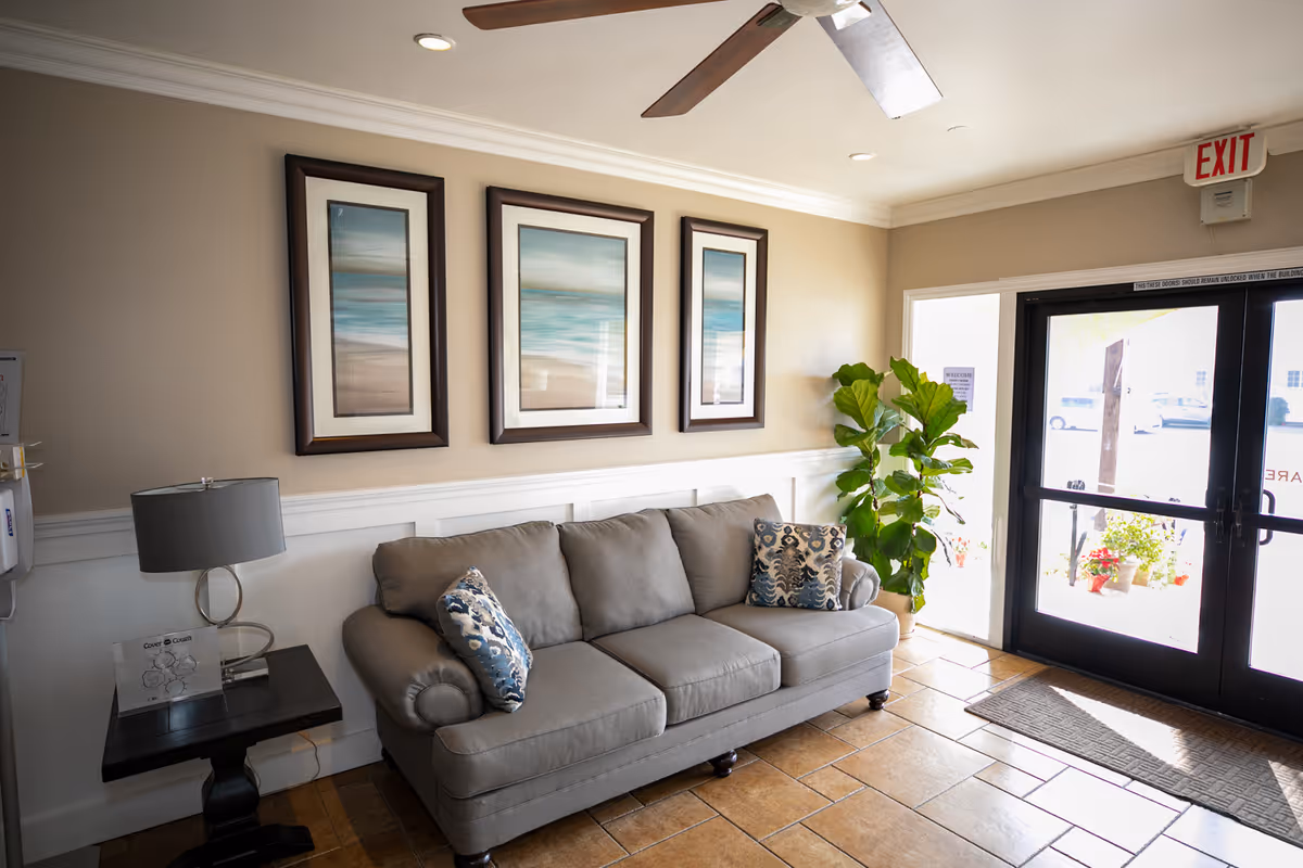 A bright lobby/lounge with a gray sofa, patterned pillows, three framed seascape prints, a side table with lamp, potted plant, and glass entry doors.