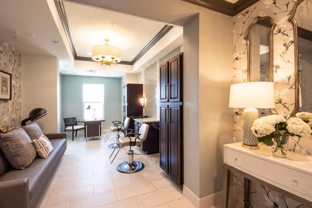 A well-lit interior room with a gray couch on the left, two salon chairs in front of a dark wood cabinet, a small desk with a chair near a window, and a white console table with a lamp and flowers on the right. The walls have floral wallpaper and the floor is tiled.