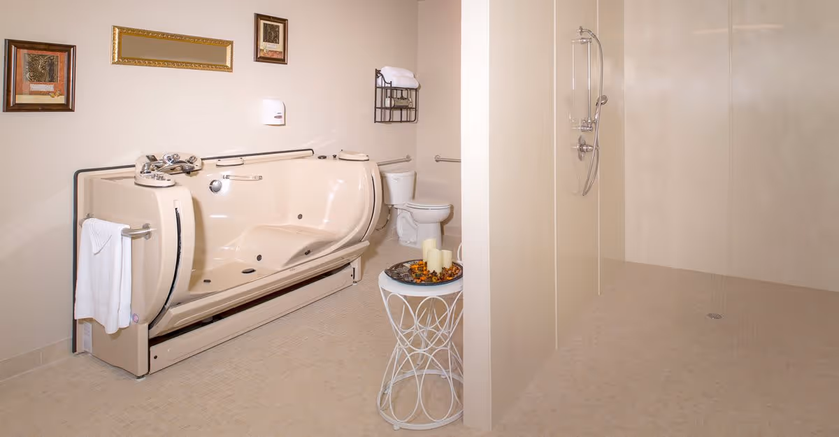 A spacious bathroom featuring a large beige walk-in bathtub with handrails and controls, a white toilet, a wall-mounted towel rack with folded towels, a small round table with decorative candles, and a walk-in shower with a handheld showerhead. The walls are light-colored and decorated with framed artwork.