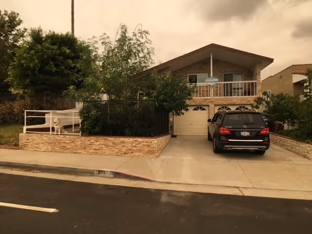 Front exterior view of a two-story residential building with a garage and a black SUV parked in the driveway. The house has a balcony with a railing and is surrounded by greenery including trees and bushes. There is a small fenced area with a white dog sitting inside it.