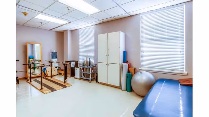 A rehabilitation room with parallel bars for walking exercises, a large mirror, a water dispenser, exercise balls, foam rollers, and a padded treatment table near two windows with blinds.