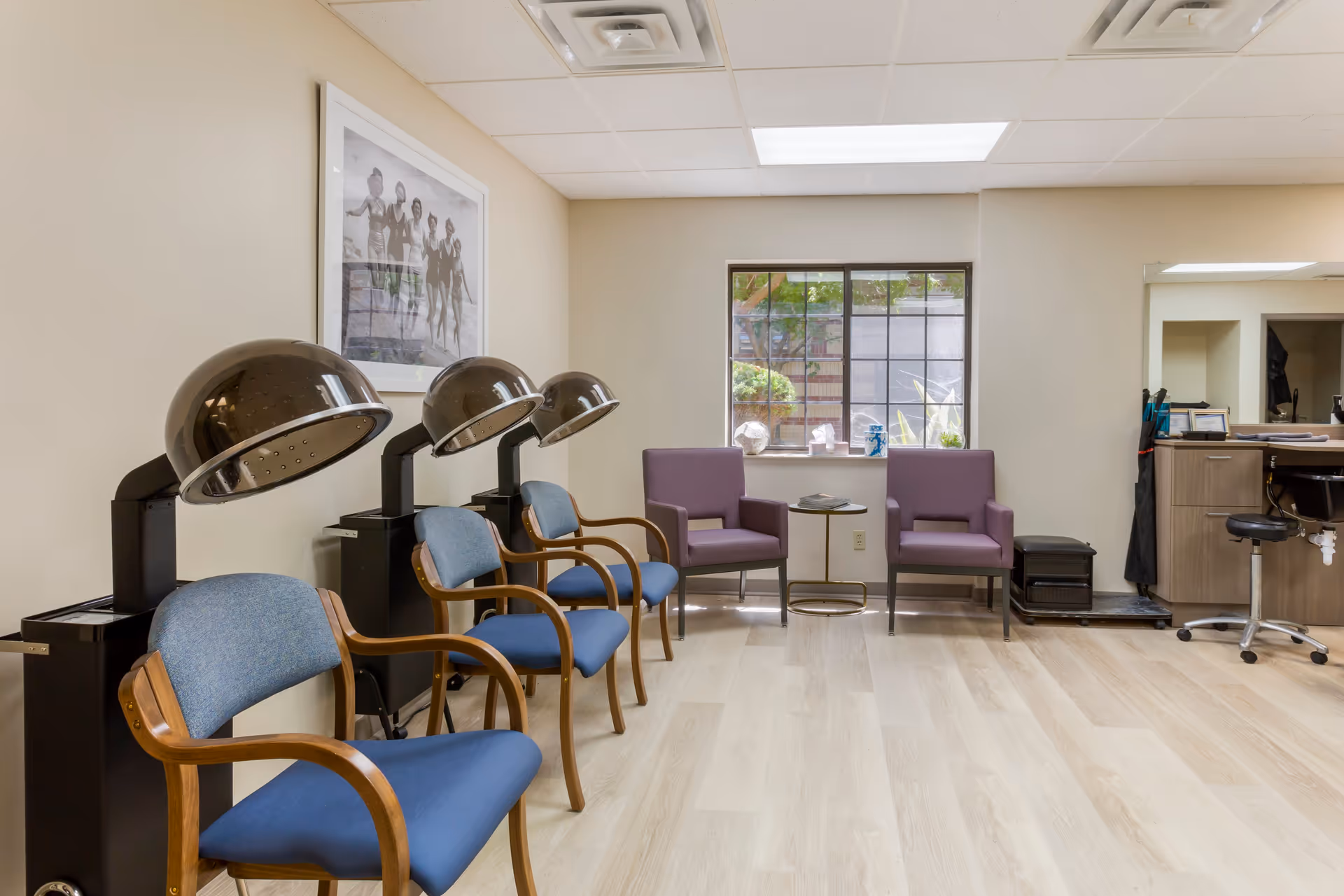 Interior view of a salon area in a senior living facility with three hair drying chairs with blue seats and wooden armrests on the left, two purple armchairs with a small round table between them near a window, and a counter with a mirror and a black swivel stool on the right.